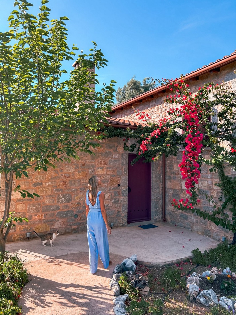 Holly in a blue jumpsuit outside a stone villa with pink bougainvillea in Greece